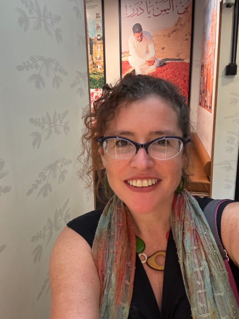 Photo of white woman with glasses and curly hair, with art that has Arabic script, in the background.
